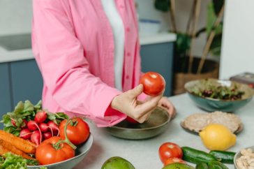 Person in pink overshirt holding a fresh tomato with various organic vegetables on a countertop.